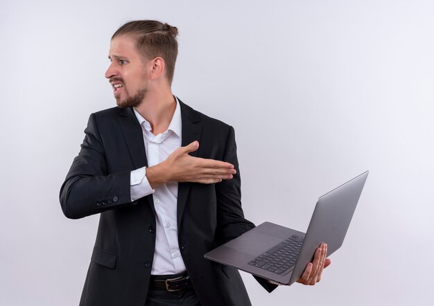 Hübscher Geschäftsmann, der Anzug hält, der Laptop-Computer hält, der verwirrt und sehr besorgt über weißem Hintergrund steht