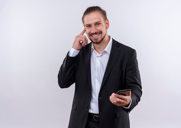 Hübscher Geschäftsmann, der Anzug hält, der Smartphone hält, das seine Schläfe zeigt, die große Idee hat, über weißem Hintergrund zu stehen
