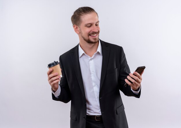 Hübscher Geschäftsmann, der Anzug hält, der Smartphone und Kaffeetasse glücklich und positiv steht über weißem Hintergrund hält