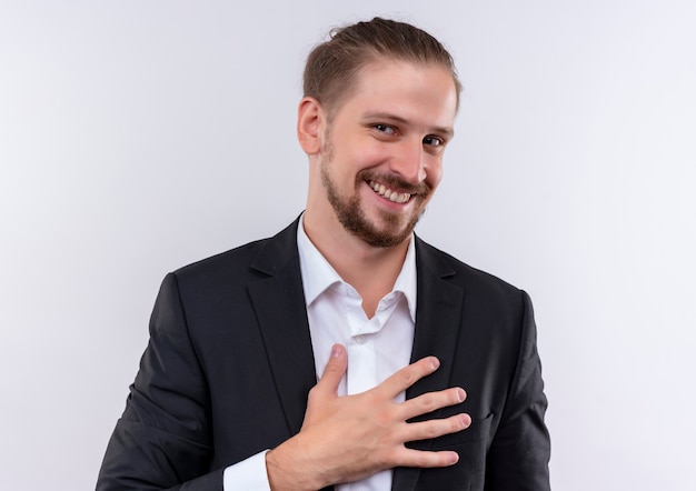 Hübscher Geschäftsmann, der Anzug hält Hand auf seiner Brust betrachtet Kamera mit Lächeln auf Gesicht, das über weißem Hintergrund steht