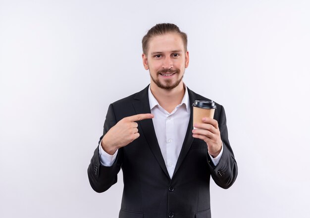 Hübscher Geschäftsmann, der Anzug hält Kaffeetasse zeigt mit dem Finger darauf lächelnd über weißem Hintergrund stehend