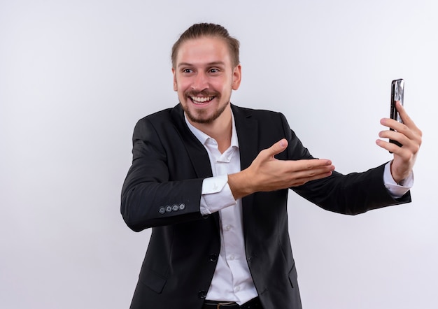 Hübscher Geschäftsmann, der Anzug hält Smartphone hält, das es mit Arm seiner Hand präsentiert, der fröhlich über weißem Hintergrund stehend lächelt