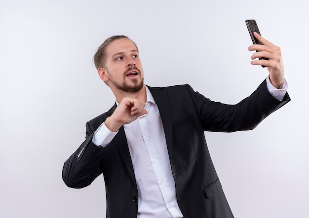 Hübscher Geschäftsmann, der Anzug hält Smartphone hält Selfie, das sicher steht, über weißem Hintergrund steht