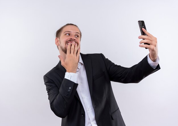 Hübscher Geschäftsmann, der Anzug hält Smartphone hält Selfie mit schüchternem Lächeln auf Gesicht steht über weißem Hintergrund