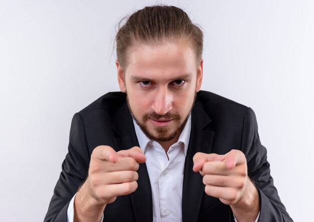 Hübscher Geschäftsmann, der Anzug trägt, der mit Zeigefingern zur Kamera mit ernstem Stehen über weißem Hintergrund zeigt