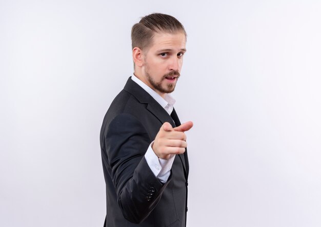 Hübscher Geschäftsmann, der Anzug trägt, der zuversichtlich zeigt, mit dem Finger zur Kamera zu stehen, die über weißem Hintergrund steht