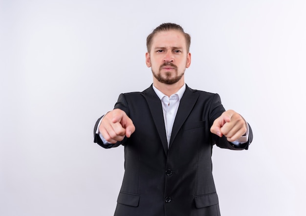 Hübscher Geschäftsmann, der Anzug trägt, der zuversichtlich zeigt, mit Zeigefingern auf Kamera zu zeigen, die über weißem Hintergrund steht