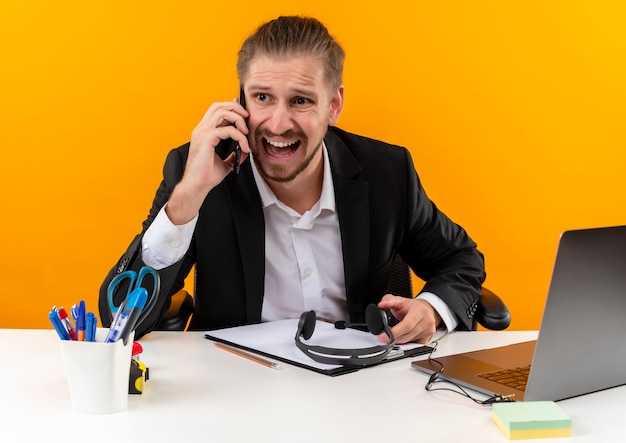 Hübscher Geschäftsmann im Anzug, der auf Laptop arbeitet, der verwirrt und unzufrieden am Tisch in offise über orange Hintergrund sitzt