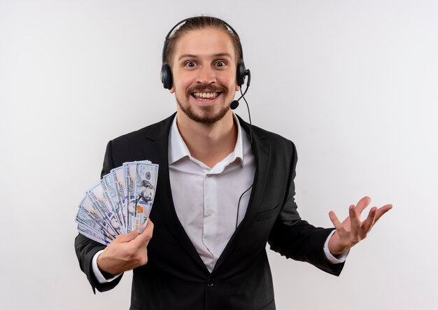 Hübscher Geschäftsmann im Anzug und in den Kopfhörern mit einem Mikrofon, das Kamera betrachtet, die Bargeld glücklich und aufgeregt über weißem Hintergrund stehend zeigt