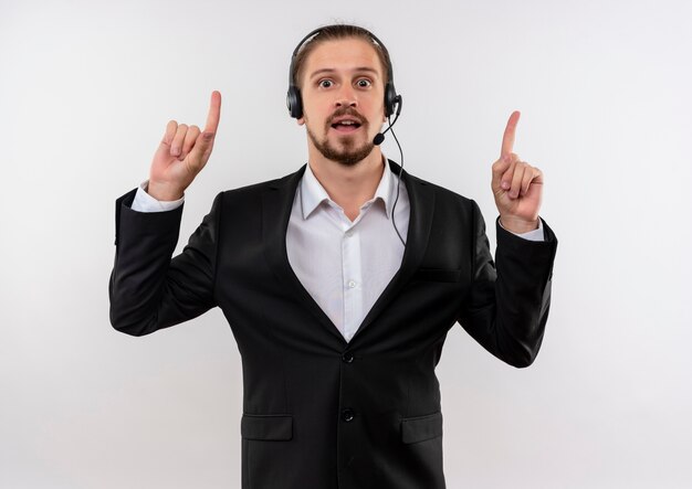 Hübscher Geschäftsmann im Anzug und in den Kopfhörern mit einem Mikrofon, das Kamera glücklich und positiv zeigt, mit Zeigefingern oben steht über weißem Hintergrund
