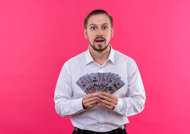 Hübscher Geschäftsmann im weißen Hemd, das Bargeld sieht, das über rosa Hintergrund überrascht steht