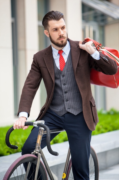 Hübscher Geschäftsmann in einer Jacke mit roter Tasche, die auf seinem Fahrrad auf Stadtstraßen sitzt.