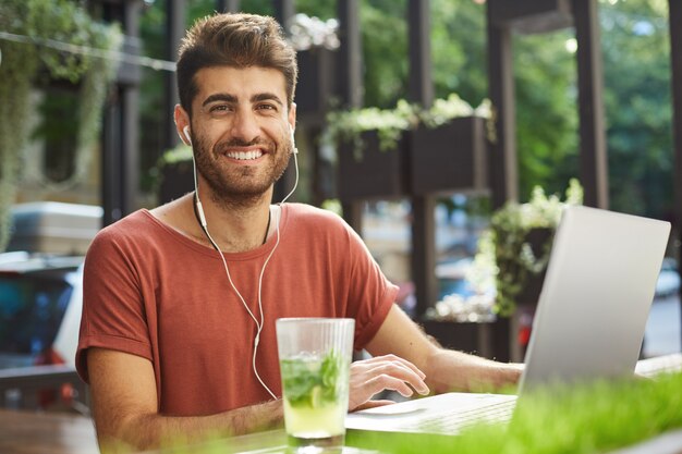 Hübscher glücklicher bärtiger Mann in den Kopfhörern, der Laptop im Straßencafé benutzt und fröhlich lächelt