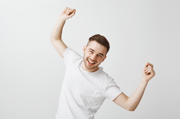 Hübscher glücklicher junger Mann, der im weißen T-Shirt über graue Wand tanzt