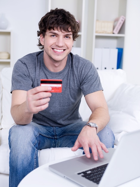 Hübscher glücklicher lächelnder Kerl, der Kreditkarte hält und Laptop verwendet - drinnen
