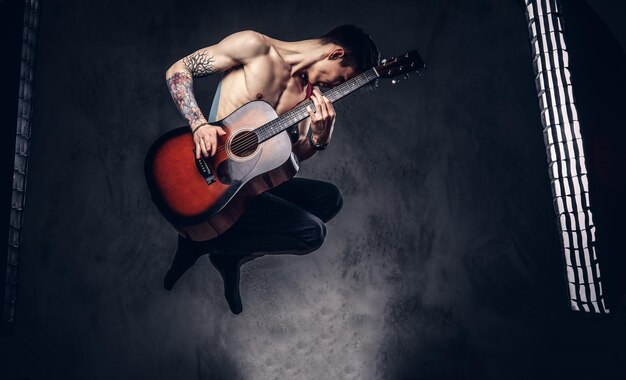 Hübscher, hemdloser junger Musiker, der beim Springen Gitarre spielt. Getrennt auf einem dunklen Hintergrund.