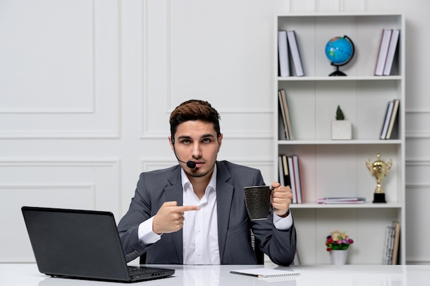 Hübscher Herr des Kundendienstes mit Computer im grauen Büroanzug, der auf Kaffeetasse zeigt