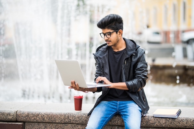 Hübscher indischer Mann mit Laptop beim Sitzen nahe dem Brunnen im Stadtzentrum an einem Tag