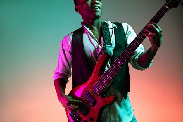 Hübscher Jazzmusiker des Afroamerikaners, der Bassgitarre im Studio auf einem Neonhintergrund spielt. Musikkonzept. Junger freudiger attraktiver Kerl, der improvisiert. Nahaufnahme Retro-Porträt.