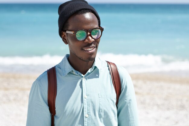 Hübscher junger afroamerikanischer Hipster, der entlang der Küste geht, gutes Wetter und Meerblick bewundert und mit dem Rücken zum weiten azurblauen Ozean steht