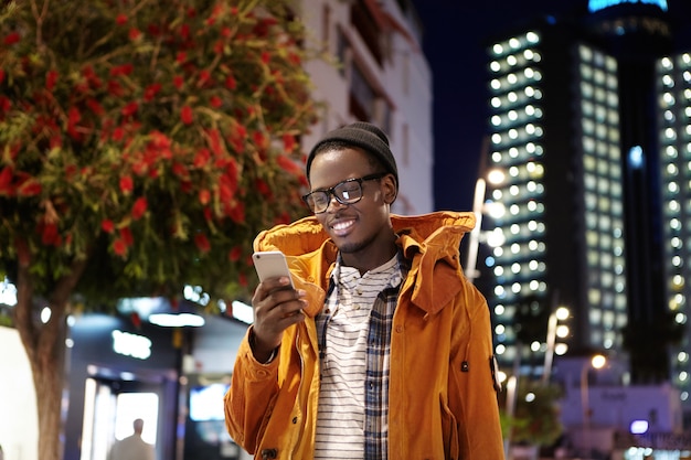 Hübscher junger dunkelhäutiger Hipster in Hut-, Brillen- und Wintermantelnachrichten auf dem Smartphone, während er nachts auf der Straße auf sie wartet und mit freudigem Lächeln auf den Bildschirm schaut