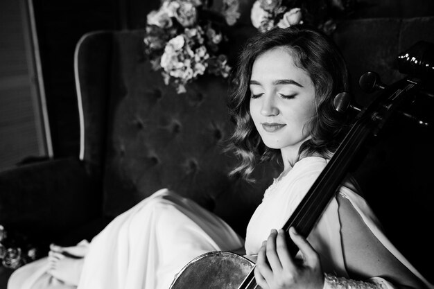 Hübscher junger Gilrl-Musiker in weißem Kleid mit Kontrabass auf braunem Vintage-Sofa