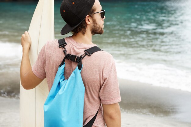 Hübscher junger kaukasischer Anfängersurfer mit Bart, der vor blauem Ozean steht und auf seinen Surflehrer wartet