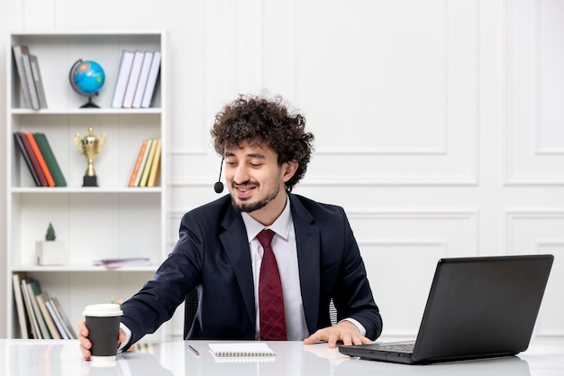 Hübscher junger Kerl des Kundendienstes im Büroanzug mit dem Laptop und dem Kopfhörer, die einen Kaffee halten