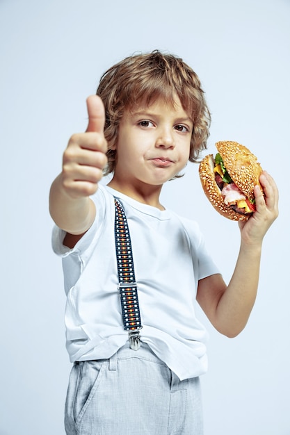 Hübscher junger lockiger Junge in Freizeitkleidung auf weißer Studiowand. Burger essen