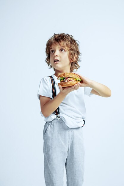 Hübscher junger lockiger Junge in Freizeitkleidung auf weißer Wand. Burger essen. Kaukasischer männlicher Vorschulkind mit hellen Gesichtsgefühlen. Kindheit, Ausdruck, Spaß haben, Fast Food. Erstaunt.