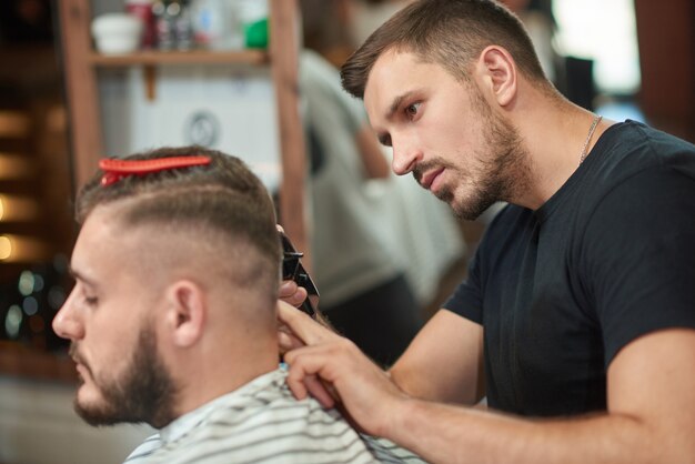 Hübscher junger männlicher Friseur, der seinem Klienten einen Haarschnitt mit einem Haarschneider gibt, der in seinem Friseursalon arbeitet.