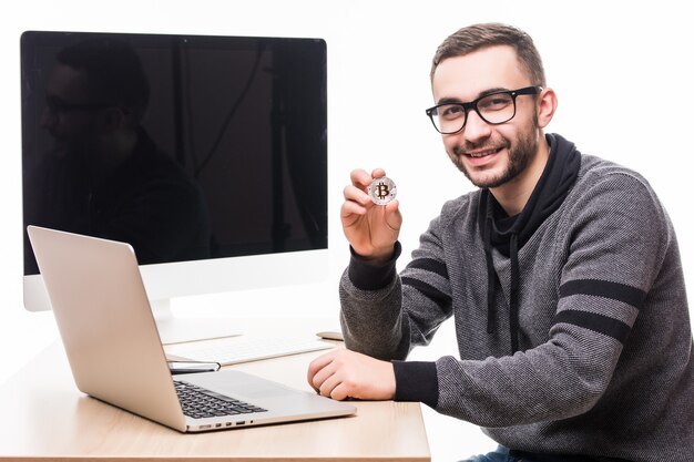 Hübscher junger Mann, der am Büroplatz mit Laptop und Bildschirm des Monitors auf seinem Rücken sitzt, zeigte Bitcoin auf Weiß