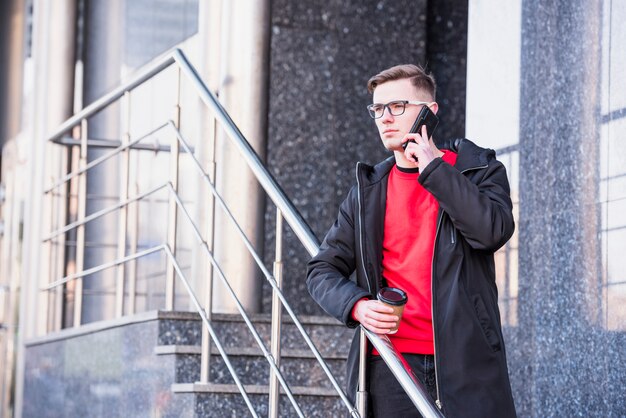 Hübscher junger Mann, der auf Skateboard am Handy hält Mitnehmerkaffeetasse steht