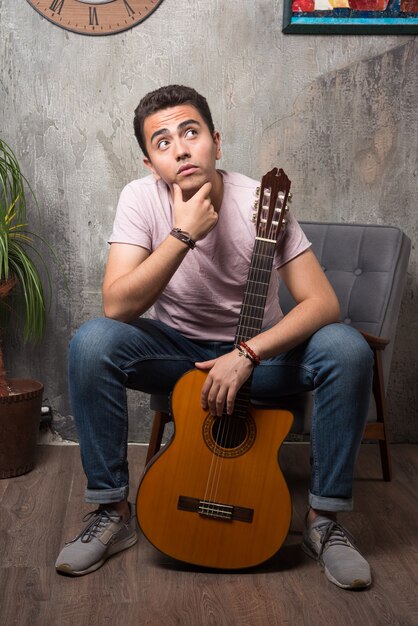 Hübscher junger Mann, der Gitarre hält und auf dem Stuhl denkt.