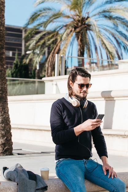 Hübscher junger Mann, der im Park mit Kaffeepapierschale unter Verwendung des Handys sitzt