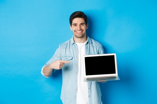 Hübscher junger Mann, der mit dem Finger auf den Bildschirm des Computers zeigt, erfreut lächelt, Promo im Internet oder auf der Website zeigt, auf blauem Hintergrund stehend.