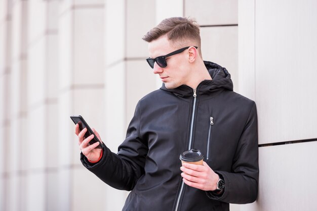 Hübscher junger Mann, der Mitnehmerkaffeetasse unter Verwendung des Smartphone hält