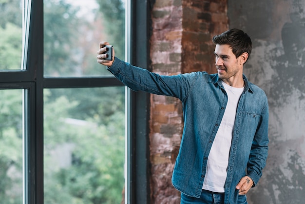 Hübscher junger Mann, der nahe dem Fenster steht, das selfie am Handy nimmt