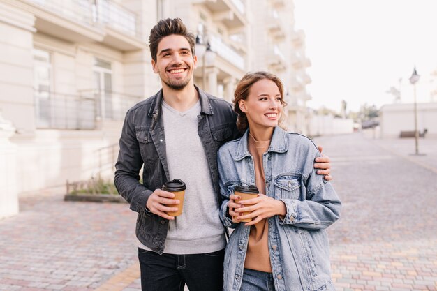 Hübscher junger Mann, der Tasse Kaffee hält und Freundin umarmt. Lächelndes Paar, das Datum im Freien genießt.