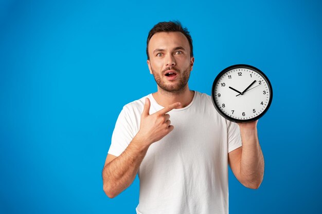 Hübscher junger Mann im weißen T-Shirt posiert mit Uhr vor blauem Hintergrund