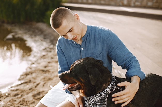 Hübscher junger Mann in lässigem Outfit, der mit süßem Hund spielt, während er in der Nähe des Sees sitzt. Junge mit blauem Hemd und weißen Jeansshorts