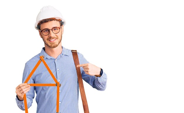 Hübscher junger Mann mit Bär und Architektenhelm, der ein Bauprojekt in der Hand hält und mit dem Finger auf sich selbst zeigt, glücklich und stolz lächelt
