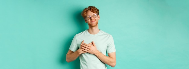 Kostenloses Foto hübscher junger mann mit roten haaren und brille, der die hände auf dem herzen hält und lächelnd sagt, danke