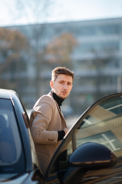 Hübscher junger Mann steht nahe am neuen modernen Auto mit geöffneter Tür am sonnigen Herbsttag