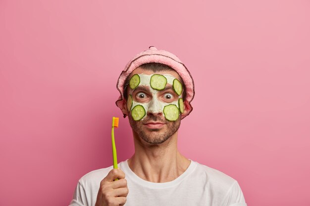 Hübscher junger Mann trägt Tonmaske mit Gurken für die Hautpflege, hält Zahnbürste, geht Zähne putzen