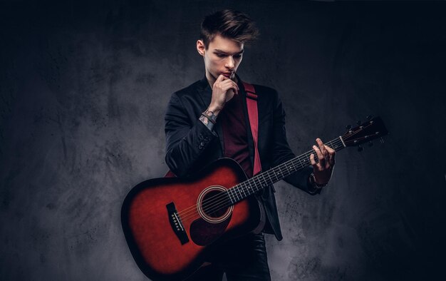 Hübscher junger nachdenklicher Musiker mit stylischem Haar in eleganter Kleidung, der mit einer Gitarre in seinen Händen posiert. Getrennt auf einem dunklen Hintergrund.