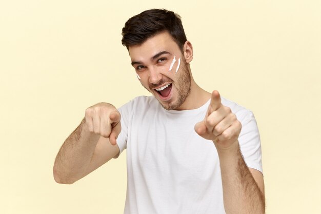Hübscher junger unrasierter Mann im weißen T-Shirt lächelnd und Zeigefinger nach vorne zeigend