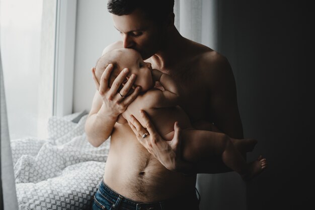Hübscher junger Vater hält neugeborenes Baby nah an seinem Herzen, das vor einem hellen Fenster steht