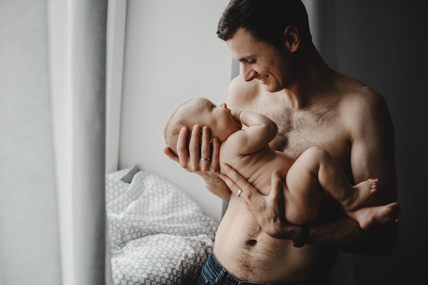 Hübscher junger Vater hält neugeborenes Baby nah an seinem Herzen, das vor einem hellen Fenster steht