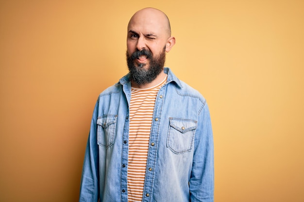 Hübscher kahlköpfiger Mann mit Bart, lässiger Jeansjacke und gestreiftem T-Shirt, der mit einem sexy Gesichtsausdruck fröhlich und glücklich in die Kamera blickt